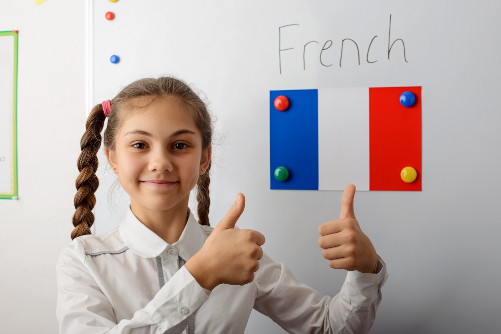 Cheerful young pupil of secondary school in white uniform with pigtails standing in front of white board and showing thumbs up